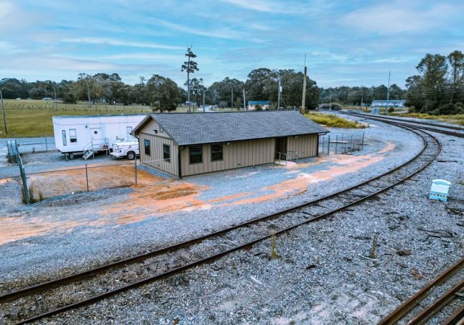 A small beige building is located next to a curved railroad track, with a white trailer and truck parked nearby. The area is surrounded by gravel, trees, and open fields beneath a cloudy sky.