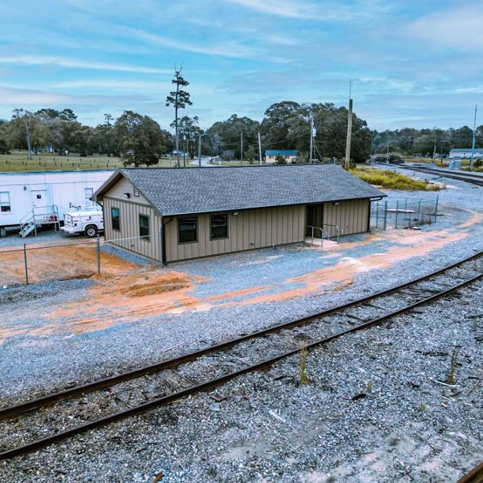 A small beige building is located next to a curved railroad track, with a white trailer and truck parked nearby. The area is surrounded by gravel, trees, and open fields beneath a cloudy sky.