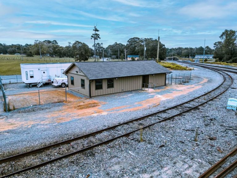 A small beige building is located next to a curved railroad track, with a white trailer and truck parked nearby. The area is surrounded by gravel, trees, and open fields beneath a cloudy sky.