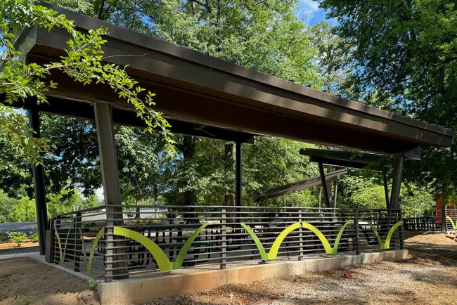 A contemporary outdoor pavilion with a slanted roof and metal railing highlighted by green accents, nestled among trees and lush greenery on a bright, sunny day.