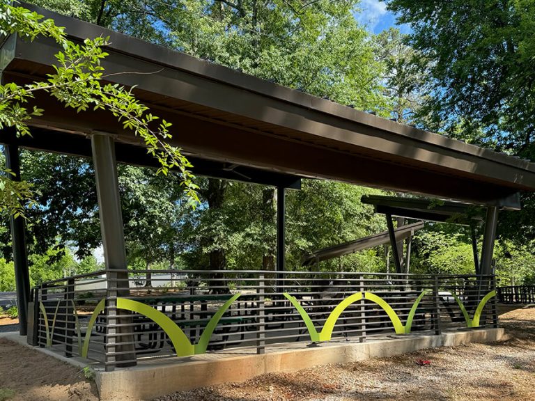 A contemporary outdoor pavilion with a slanted roof and metal railing highlighted by green accents, nestled among trees and lush greenery on a bright, sunny day.