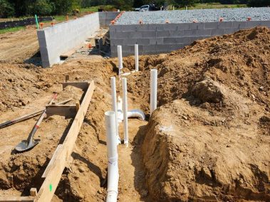 PVC pipes for plumbing are installed in the dirt at a construction site. A partially built concrete block foundation and a gravel area can be seen in the background under a blue sky.