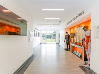 A brightly lit hallway with a large TV on the left and two mannequins in baseball uniforms on the right, near an orange display labeled MU and DIANE OWENS LOBBY. Glass doors reveal an outdoor field ahead.