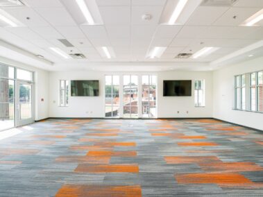 Bright, empty room with large windows, two wall-mounted TVs, and patterned carpet in gray and orange. Ceiling lights are on, and sunlight streams in from the windows, revealing a view of greenery outside.