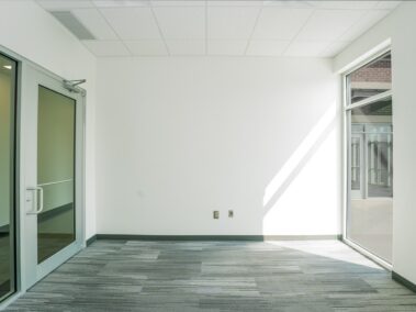 An empty office room featuring glass doors and windows, white walls, a gray patterned carpet, and sunlight streaming in from a window on the right.