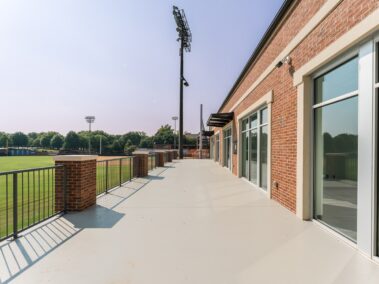 Spacious outdoor balcony featuring brick walls and large glass doors, offering a view of a grassy athletic field bordered by trees and illuminated stadium lights.