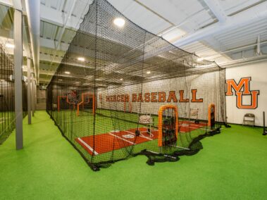 Indoor baseball training area with green turf, batting cage, and orange Mercer Baseball lettering on the wall. Two orange baseball practice nets and equipment are visible inside the cage.