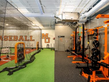Indoor baseball training facility with a batting cage on the left and orange and black weightlifting equipment on the right. BASEBALL and MU logos are visible on the back wall. Bright lighting illuminates the space.