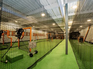 Indoor baseball training facility featuring artificial turf, several batting cages, and multiple pitching machines. The room is surrounded by netting with ball carts, protective screens, and various training equipment arranged throughout the space.