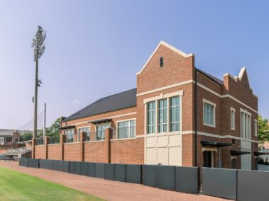 A brick building with large windows is situated beside a sports field beneath a clear blue sky. A tall light pole stands close to the field, which is bordered by a black fence.