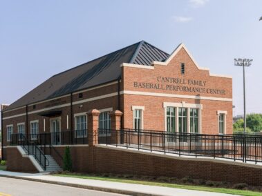 Exterior view of the red brick Cantrell Family Baseball Performance Center on a sunny day, featuring a dark metal roof, a wheelchair-accessible ramp, stairs leading to the entrance, and tall stadium lights nearby under a clear blue sky.