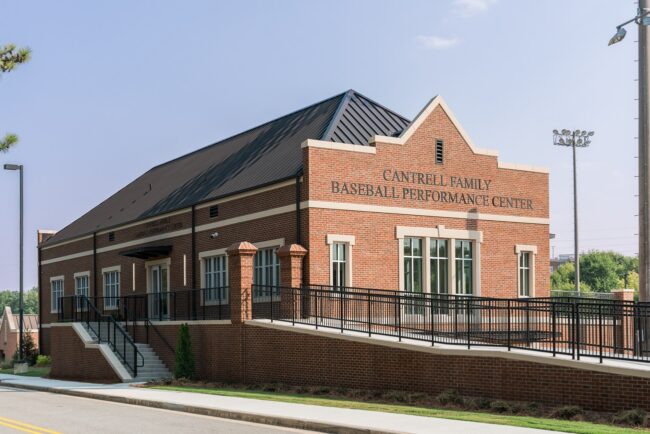 Exterior view of the red brick Cantrell Family Baseball Performance Center on a sunny day, featuring a dark metal roof, a wheelchair-accessible ramp, stairs leading to the entrance, and tall stadium lights nearby under a clear blue sky.