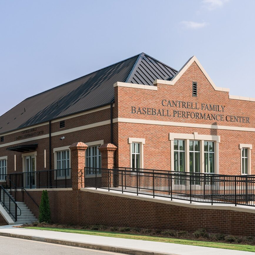 TCM07917 Exterior view of the red brick Cantrell Family Baseball Performance Center on a sunny day, featuring a dark metal roof, a wheelchair-accessible ramp, stairs leading to the entrance, and tall stadium lights nearby under a clear blue sky.