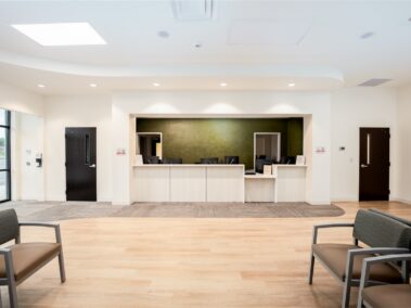 A modern, well-lit reception area features a green accent wall, several desks with monitors, and chairs arranged in the waiting area. Large windows fill the space with natural light. The environment looks clean and welcoming.
