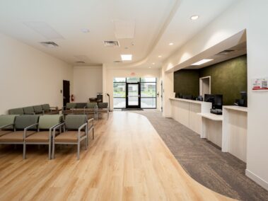 A modern, empty waiting room featuring rows of green chairs, a wooden floor, a large reception counter on the right, white walls, and double glass doors at the back allowing natural light to enter.