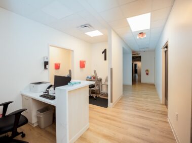 A clean medical office hallway with wood flooring, white walls, a reception desk with computer, chairs, and doors to exam rooms. A large number 1 is on the wall, and an exit sign hangs from the ceiling.
