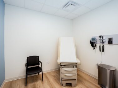 A small medical exam room featuring a patient table, a black chair, medical instruments mounted on the wall, and a trash bin. The walls are painted white and blue, with light wood flooring.