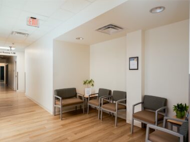 A small medical waiting area with five chairs, two plants, and a sign on a table. The hallway leads to exam rooms. Walls are white, and flooring is light wood. Overhead lights and an exit sign are visible.
