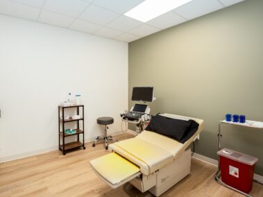 A medical exam room featuring a yellow patient chair, an ultrasound machine, a stool, a supply cart, and a sharps container. The walls are painted white and green, and the floor has a wood finish.