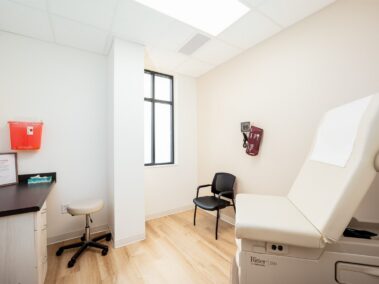 A clean and empty medical exam room featuring an exam table, a stool, a black chair, blood pressure monitor, sharps container, window, and light wood flooring brightly lit.