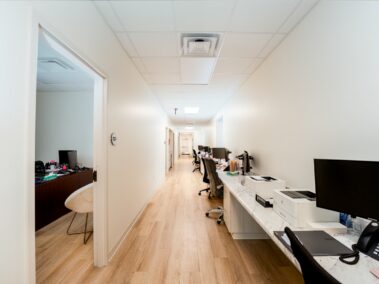 Bright office hallway featuring hardwood floors, desks equipped with computers and office supplies lined up along the right wall, and an open door on the left that leads to another office.
