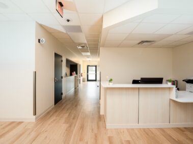 A clean, modern hallway in a medical office with light wood floors, beige walls, and a white reception desk that holds office supplies. A printer sits on the counter, and a door is visible at the end of the hall.