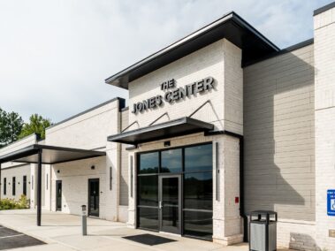Modern building with The Jones Center sign above glass entrance, accessible ramp, and handicap parking sign in front. Light brick exterior and black awnings, with trees visible in the background.