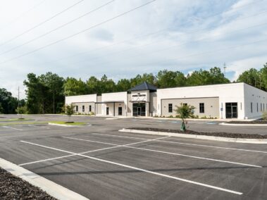 A modern one-story white building with a sign above its entrance, surrounded by trees and an empty, freshly paved parking lot beneath a partly cloudy sky.