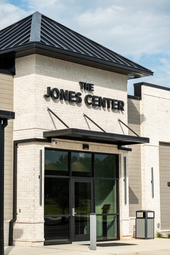 Modern building exterior with a white brick facade and black trim. Bold black letters read The Jones Center above a glass door with the number 285. Trash cans and a small plant appear nearby.