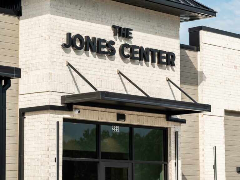 Modern building exterior with a white brick facade and black trim. Bold black letters read The Jones Center above a glass door with the number 285. Trash cans and a small plant appear nearby.