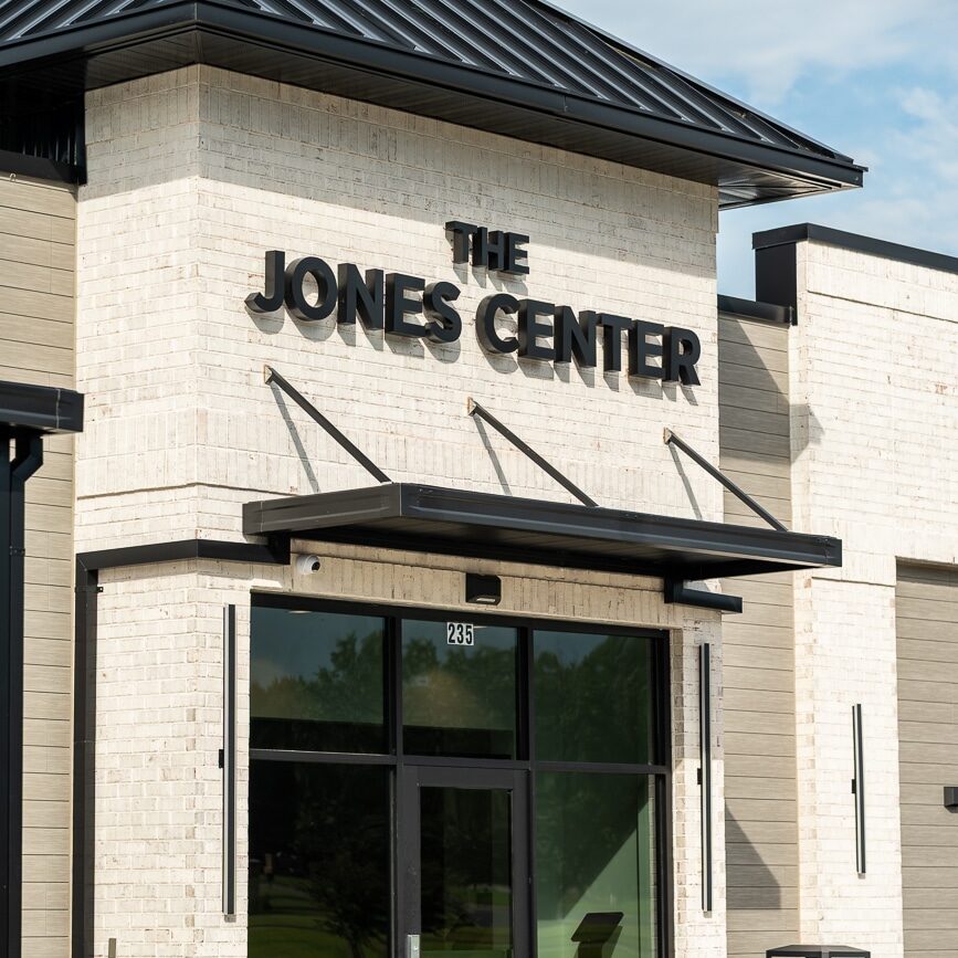 TCM08081 Modern building exterior with a white brick facade and black trim. Bold black letters read The Jones Center above a glass door with the number 285. Trash cans and a small plant appear nearby.